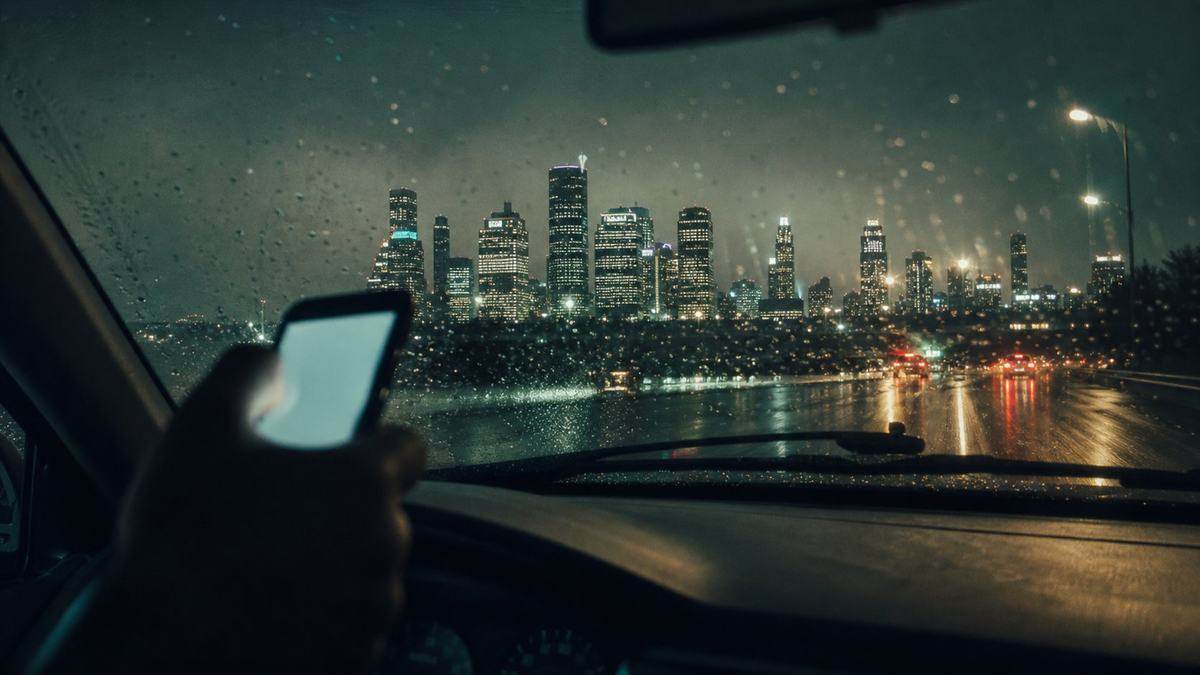 A hand holding a glowing phone inside a car on a wet road, the Houston skyline ahead